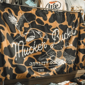 The Wood Ducks Duck Camo Flag by Mucker Bucket features a brown and black duck camo pattern with two flying ducks and the text “Mucker Bucket Quality Goods TRD MRK” printed on it. Made from durable polyester, it includes metal grommets for easy hanging.