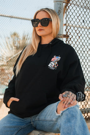 A woman with long blonde hair and black sunglasses wears the Relentless Betrayal Revolution Premium Hoodie, featuring a colorful skull graphic, paired with ripped blue jeans, as she sits outdoors against a chain-link fence.