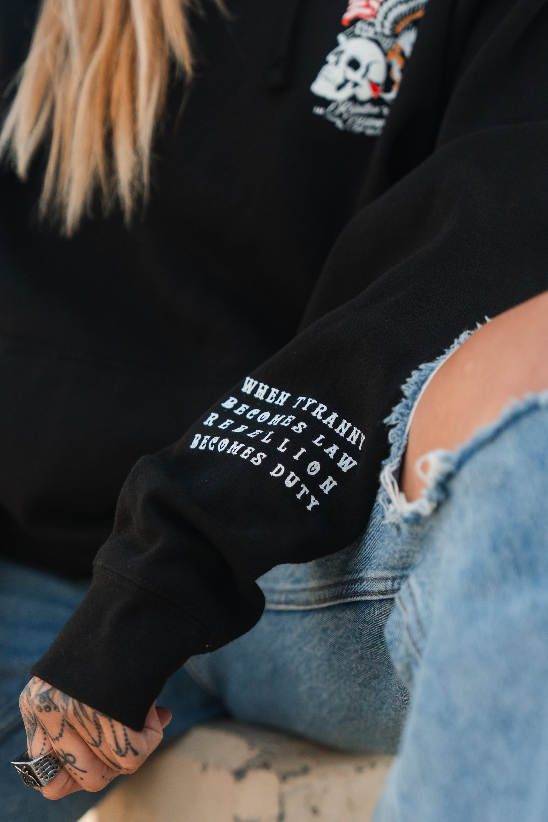 A person with long blonde hair wears the Relentless Betrayal Revolution Premium Hoodie, featuring a skull graphic and an arm patch reading "When tyranny becomes law, rebellion becomes duty." They pair it with ripped blue jeans and show off hand tattoos.