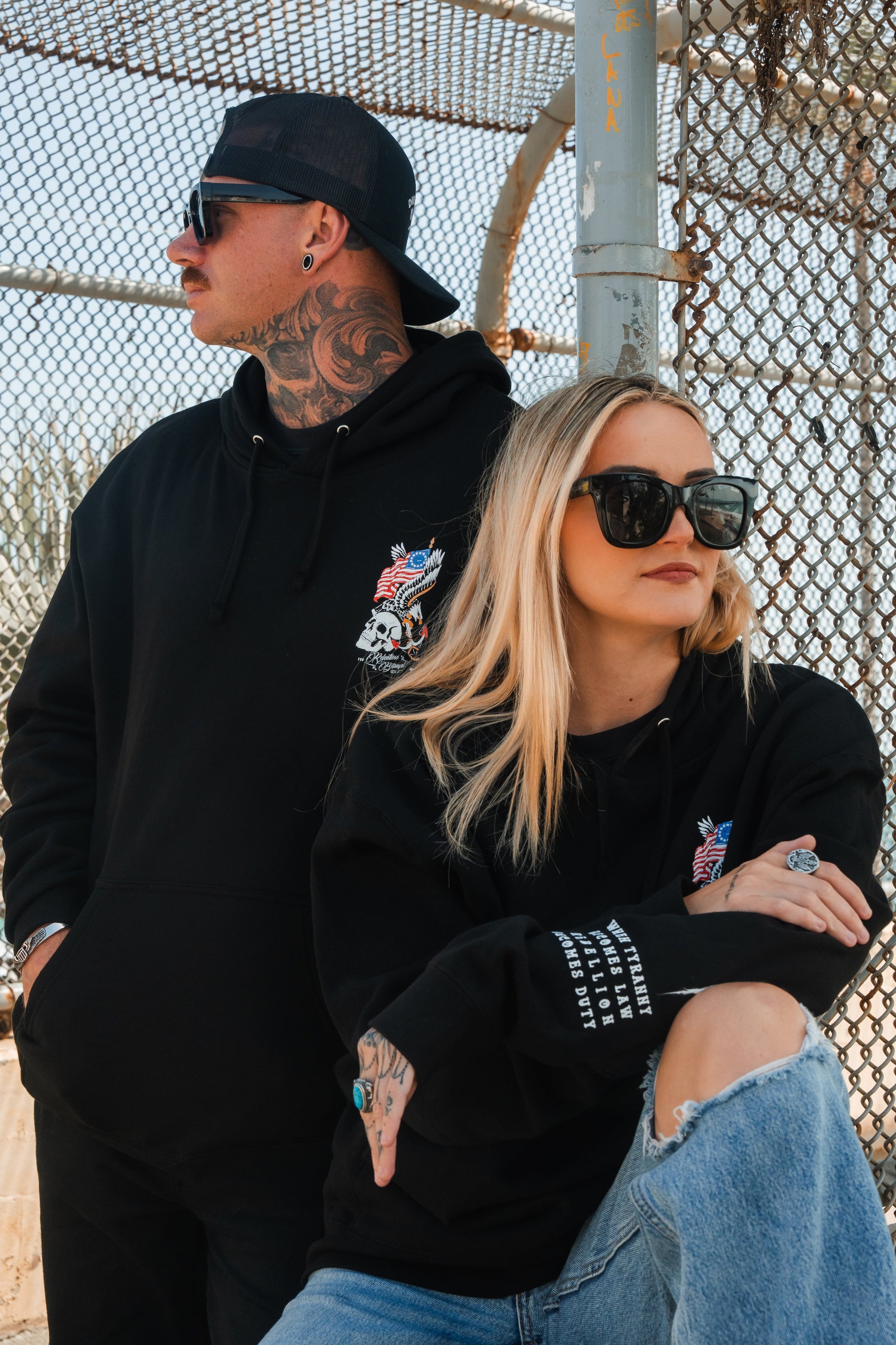 Two people in Relentless Betrayal's Revolution Premium Hoodies and sunglasses pose by a chain-link fence. The woman, in ripped jeans, sits arms crossed; the man stands behind her, cap backward, confidently showing his tattoos.