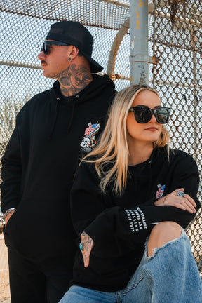 Two people in Relentless Betrayal's Revolution Premium Hoodies and sunglasses pose by a chain-link fence. The woman, in ripped jeans, sits arms crossed; the man stands behind her, cap backward, confidently showing his tattoos.