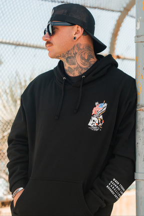 A tattooed man in a Relentless Betrayal Revolution Premium Hoodie, sunglasses, and a backward cap leans against a pole by a chain-link fence outdoors.
