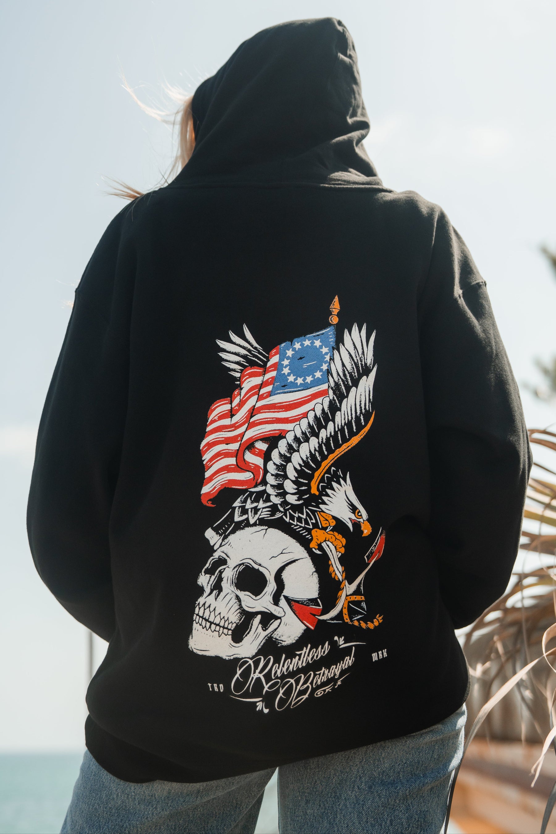 Someone wears the Revolution Premium Hoodie by Relentless Betrayal, featuring a bold skull, eagle, and waving American flag with white and red text over a beach scene—showcasing integrity and standout style.