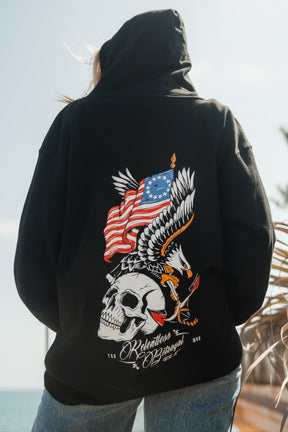 Someone wears the Revolution Premium Hoodie by Relentless Betrayal, featuring a bold skull, eagle, and waving American flag with white and red text over a beach scene—showcasing integrity and standout style.