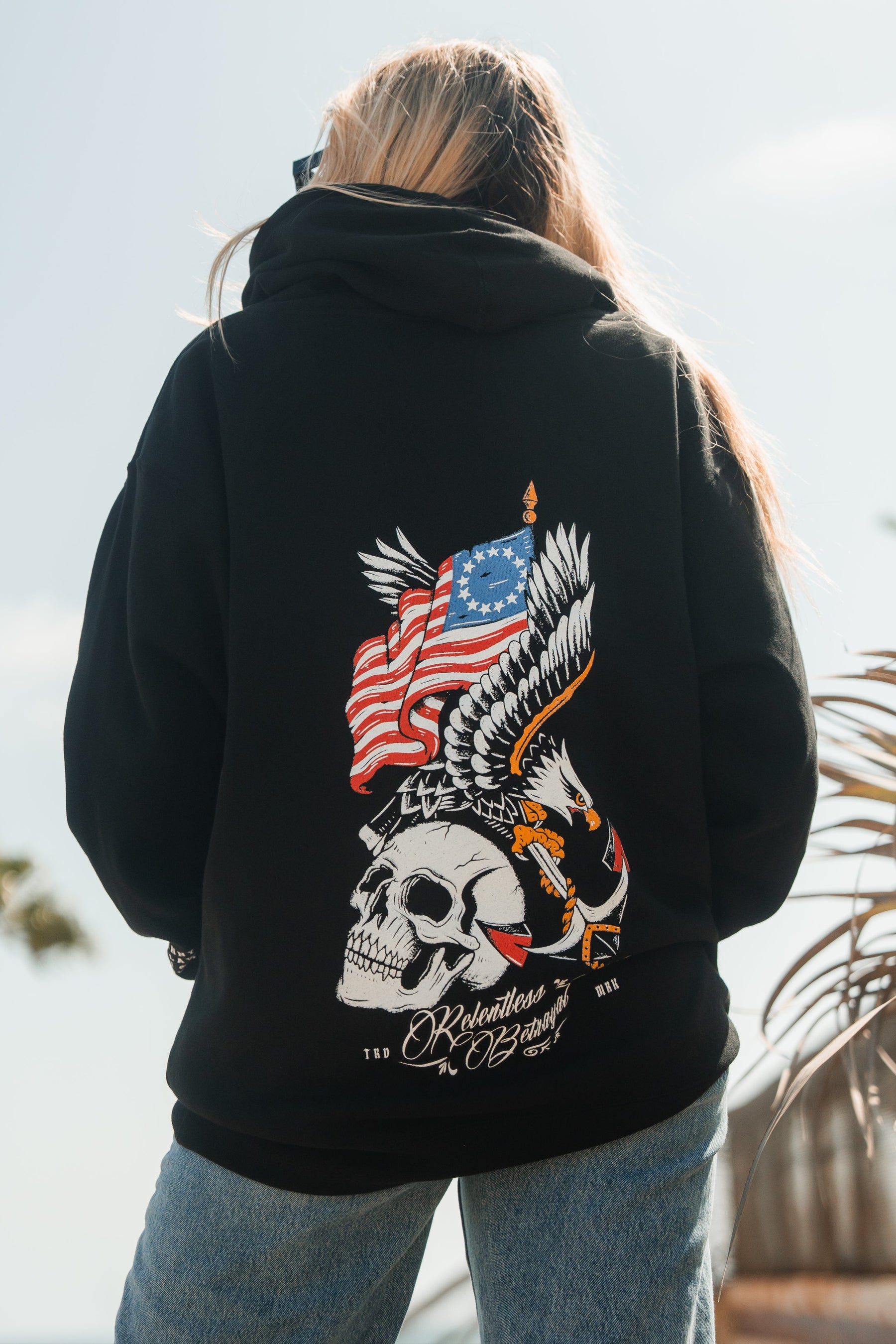 Outdoors in sunlight, a person faces away from the camera wearing the Relentless Betrayal Revolution Premium Hoodie—a black statement piece with a bold back graphic featuring a skull, eagle, and American flag.