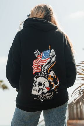 Outdoors in sunlight, a person faces away from the camera wearing the Relentless Betrayal Revolution Premium Hoodie—a black statement piece with a bold back graphic featuring a skull, eagle, and American flag.