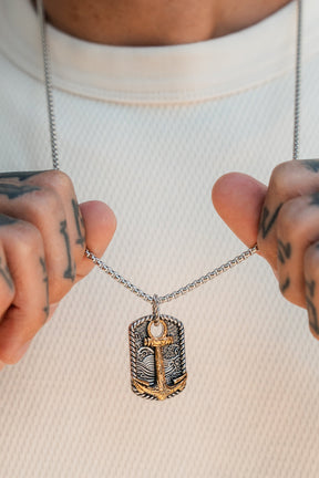 Hands holding the Relentless Betrayal Beauty in the Storm Necklace, featuring a stainless steel rectangular pendant with an engraved gold anchor design, against a white textured shirt.