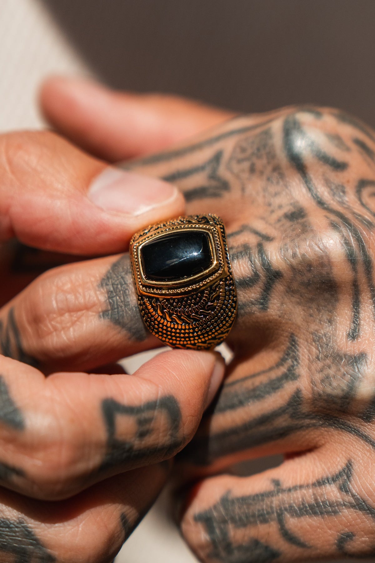 A tattooed hand holds The Aztec GOLD Ring by Relentless Betrayal, a handcrafted piece with an ornate design and a striking large black rectangular stone.