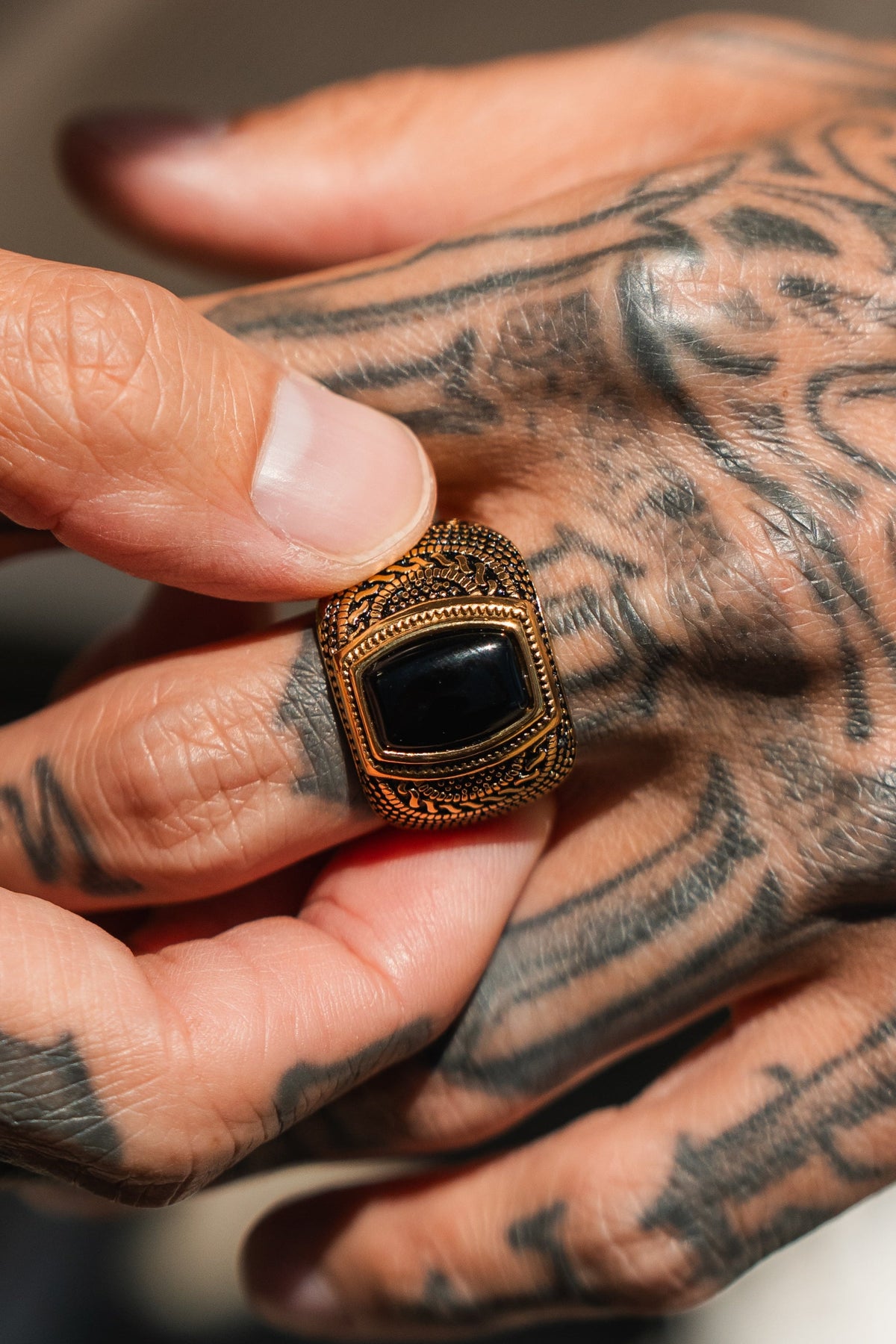 A hand adorned with black tattoos holds The Aztec GOLD Ring by Relentless Betrayal—a handcrafted piece featuring a striking, large rectangular black stone—between the thumb and index finger.