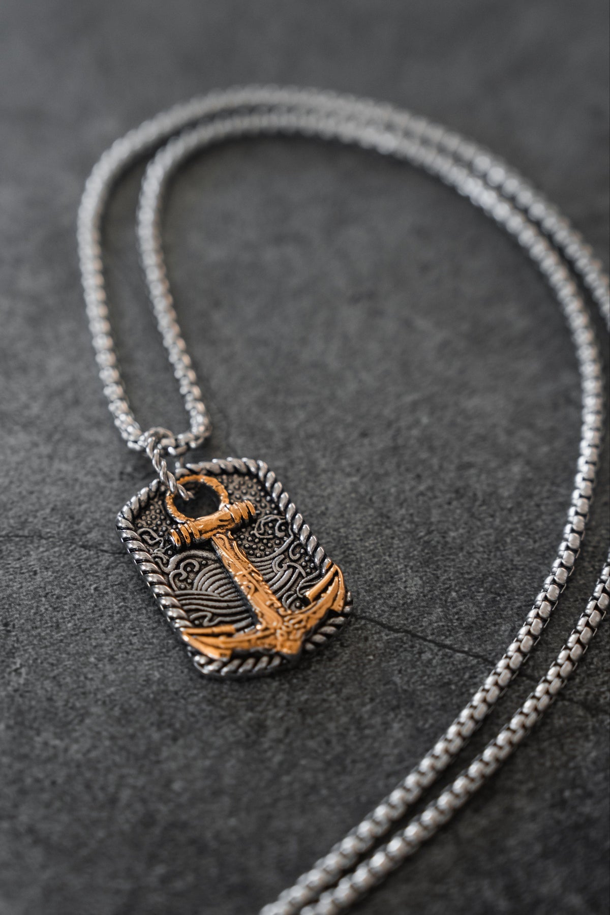 Introducing the Beauty in the Storm Necklace by Relentless Betrayal—a handcrafted silver necklace featuring a rectangular pendant adorned with an ornate gold anchor design, beautifully showcased on a dark textured background. This elegant piece of jewelry adds sophistication to any ensemble.