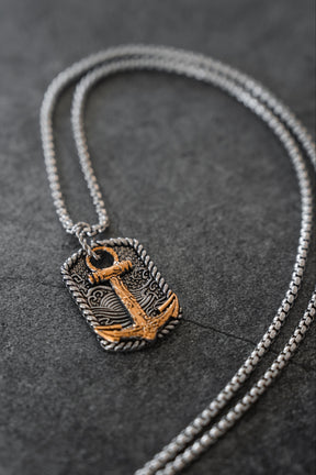 Introducing the Beauty in the Storm Necklace by Relentless Betrayal—a handcrafted silver necklace featuring a rectangular pendant adorned with an ornate gold anchor design, beautifully showcased on a dark textured background. This elegant piece of jewelry adds sophistication to any ensemble.