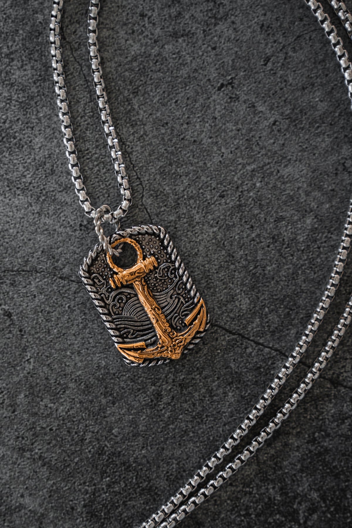 The Beauty in the Storm Necklace by Relentless Betrayal features a handcrafted silver chain and a rectangular pendant adorned with an ornate gold anchor design, elegantly displayed on a dark textured surface.