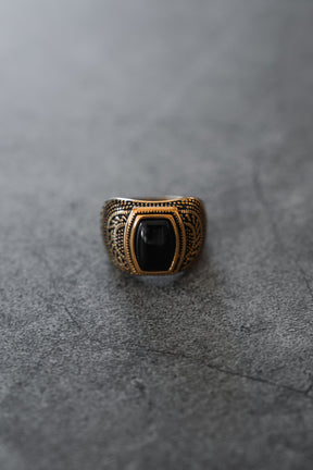 A close-up of The Aztec GOLD Ring by Relentless Betrayal, showcasing a striking black stone set in an ornate gold and silver band, displayed on a gray textured surface.