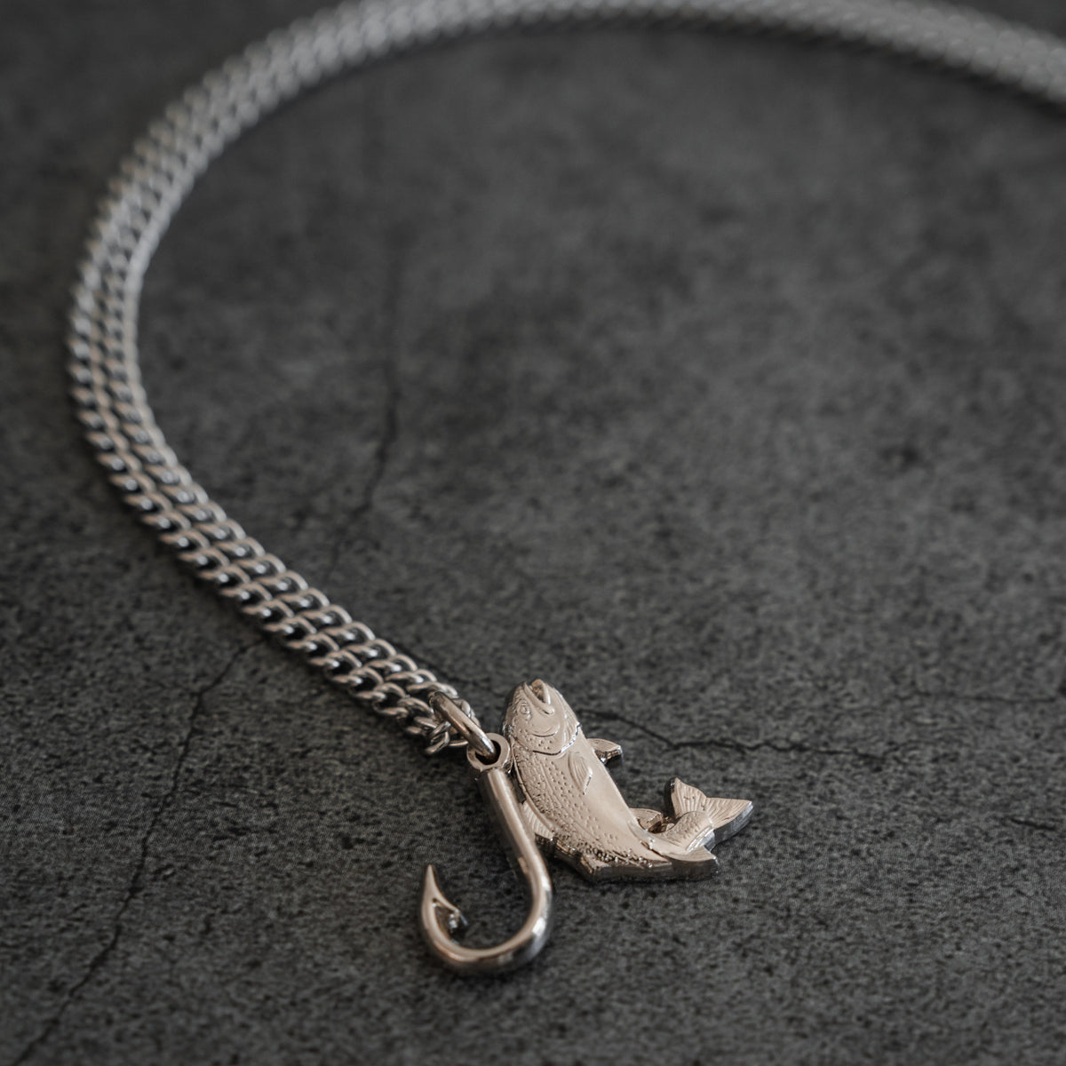 The Mucker Bucket Fly Fishin’ Trout Hunter – Stainless Steel Necklace, featuring a silver trout and hook pendant, rests on a textured gray surface, suspended from a durable stainless steel chain.