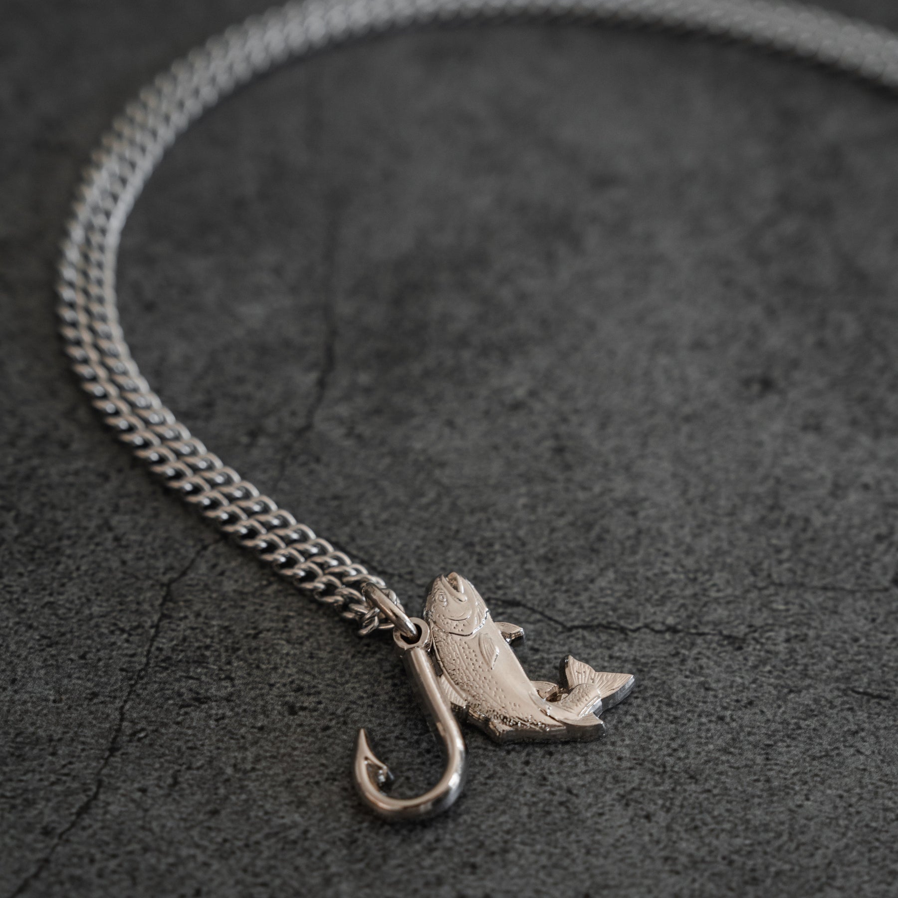 The Mucker Bucket Fly Fishin’ Trout Hunter – Stainless Steel Necklace, featuring a silver trout and hook pendant, rests on a textured gray surface, suspended from a durable stainless steel chain.
