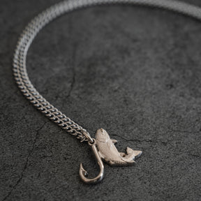 The Mucker Bucket Fly Fishin’ Trout Hunter – Stainless Steel Necklace, featuring a silver trout and hook pendant, rests on a textured gray surface, suspended from a durable stainless steel chain.