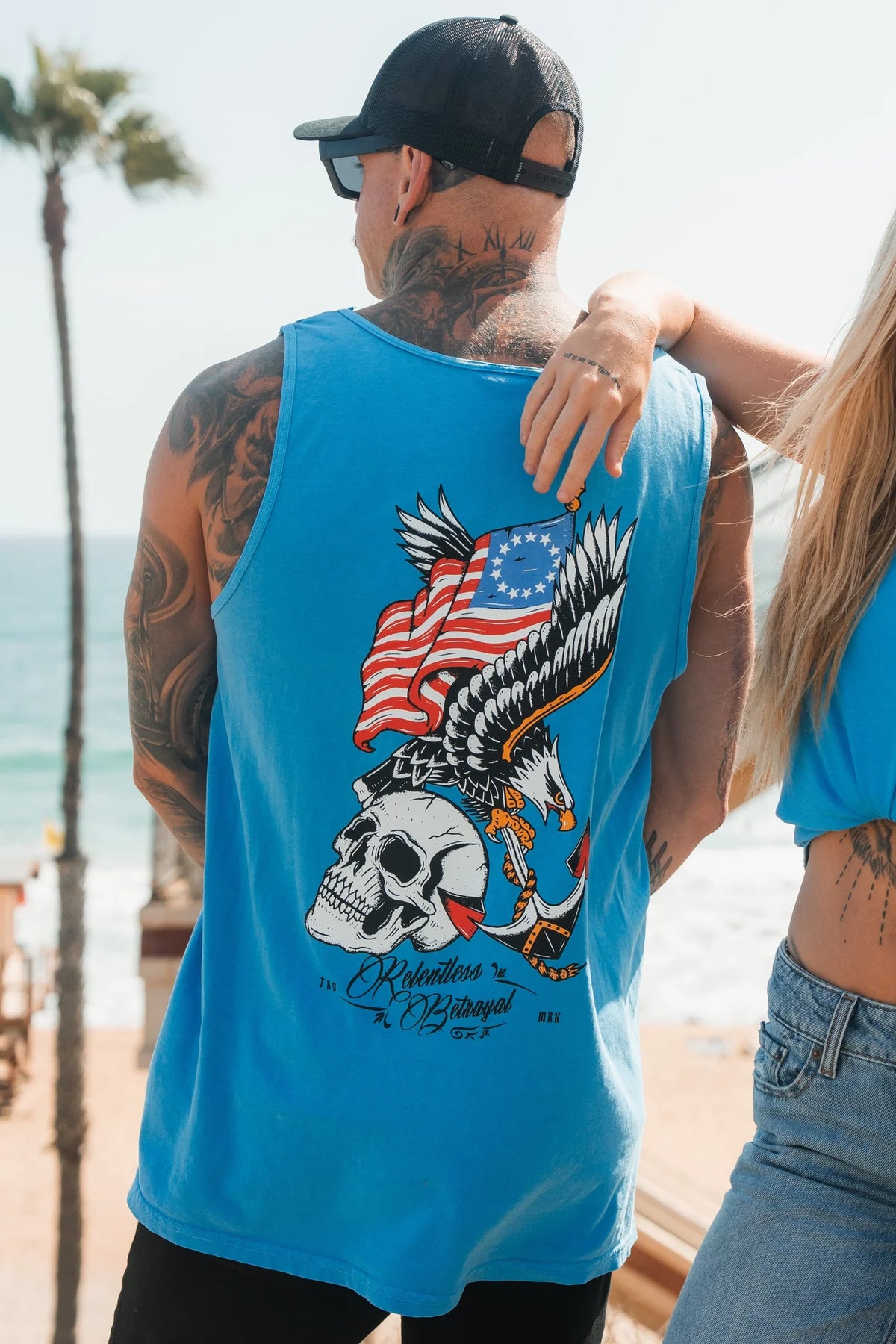 A tattooed man in a Relentless Betrayal Revolution Premium Tank-Blue with eagle, skull, and flag graphic stands by the beach next to a woman in a matching top, with palm trees and the ocean in the background.