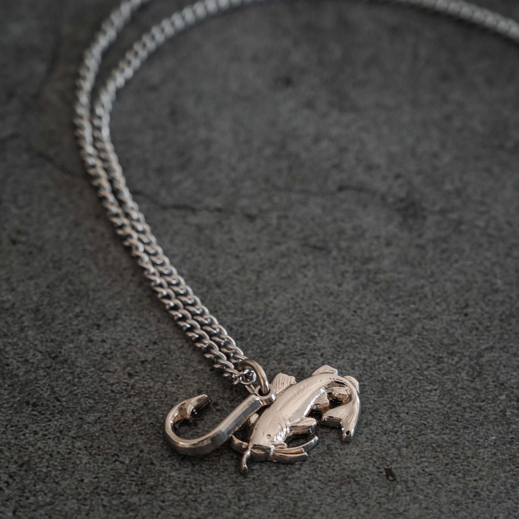 The Mud Cat Noodler Stainless Steel Necklace by Mucker Bucket features a silver chain with a fish and fish hook pendant, crafted in the USA and shown on a dark grey stone surface.