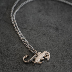 The Mud Cat Noodler Stainless Steel Necklace by Mucker Bucket features a silver chain with a fish and fish hook pendant, crafted in the USA and shown on a dark grey stone surface.
