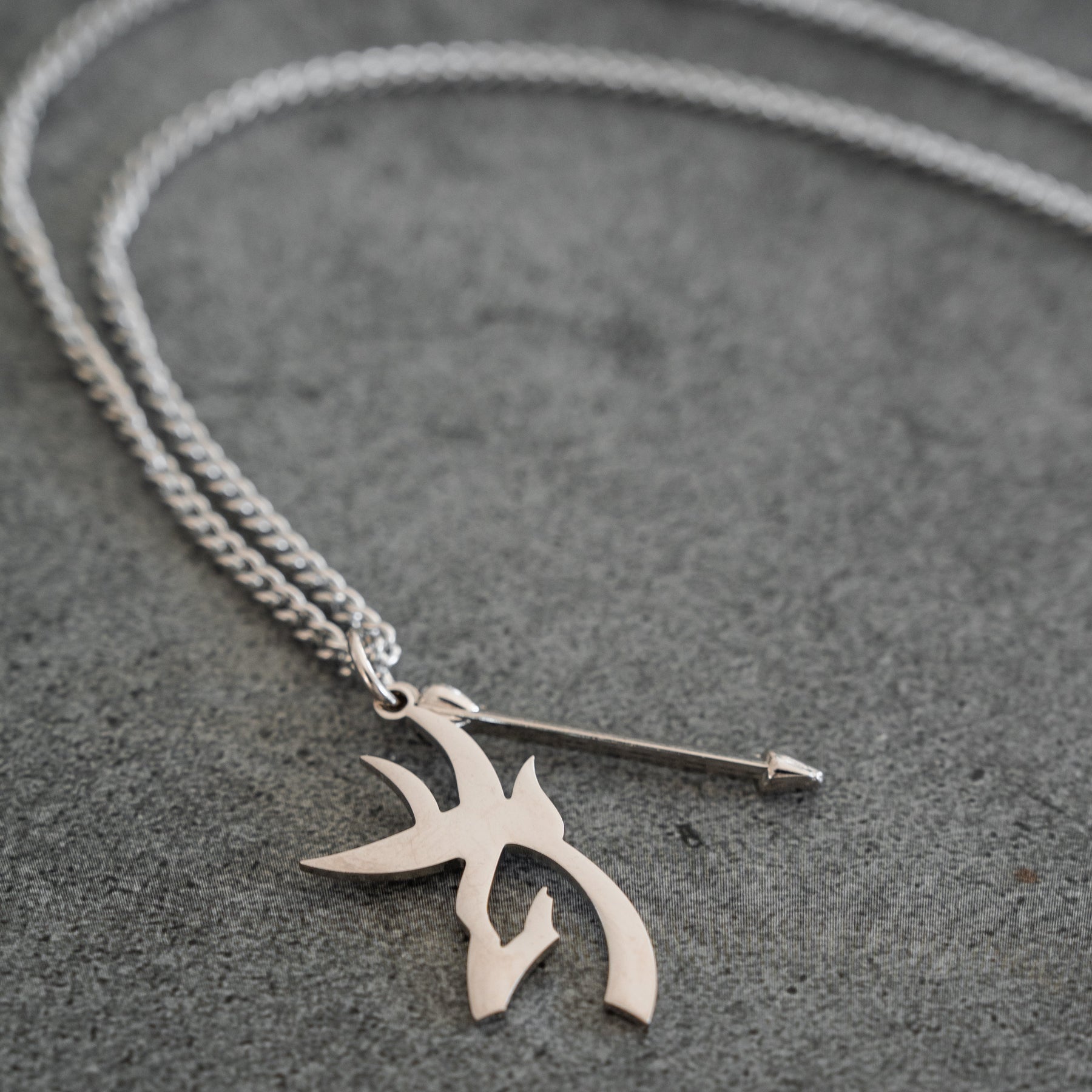 The Mucker Bucket Big Buck Bow Hunter - Stainless Steel Necklace, featuring a chain link with two pendant charms—one shaped like an arrow and the other adorned with an abstract, tribal-inspired design—rests on a gray surface. This distinctive piece includes a stainless steel pendant that celebrates the spirit of the Bow Hunter and is proudly made in the USA.