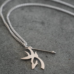The Mucker Bucket Big Buck Bow Hunter - Stainless Steel Necklace, featuring a chain link with two pendant charms—one shaped like an arrow and the other adorned with an abstract, tribal-inspired design—rests on a gray surface. This distinctive piece includes a stainless steel pendant that celebrates the spirit of the Bow Hunter and is proudly made in the USA.