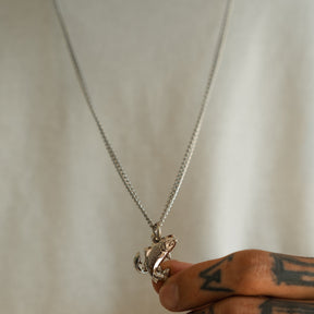A tattooed hand holds the Mucker Bucket Fly Fishin’ Trout Hunter Stainless Steel Necklace, featuring a detailed trout pendant on a stainless steel chain, with a white shirt visible in the background.