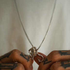 A person with tattooed fingers holds the Mud Cat Noodler - Stainless Steel Necklace by Mucker Bucket, which features a stainless steel fish and hook pendant suspended from a matching chain, displayed against a plain white shirt background.