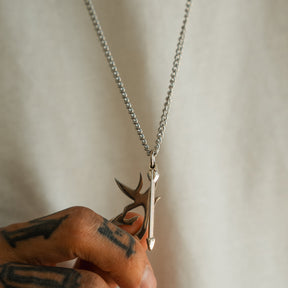 A hand with tattoos holds the Mucker Bucket Big Buck Bow Hunter Stainless Steel Necklace, featuring a silver pendant with a stylized arrow and abstract design inspired by the spirit of the bow hunter, set against a plain light background.