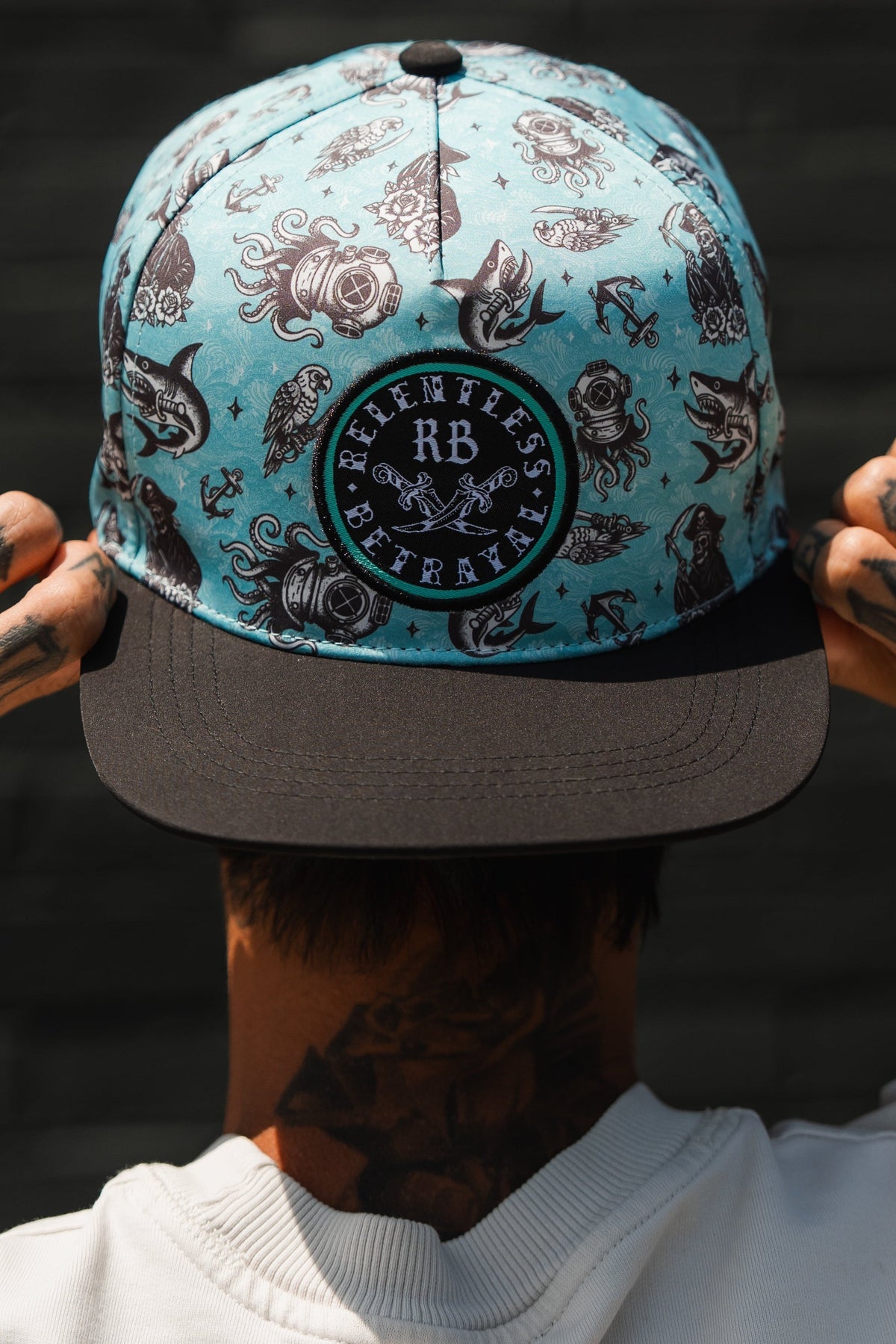 A person with neck and hand tattoos, facing away, wears the Relentless Betrayal Sea Swallows the Weak PVC Snapback—a teal and black water-resistant hat featuring sea creature designs and a circular patch.