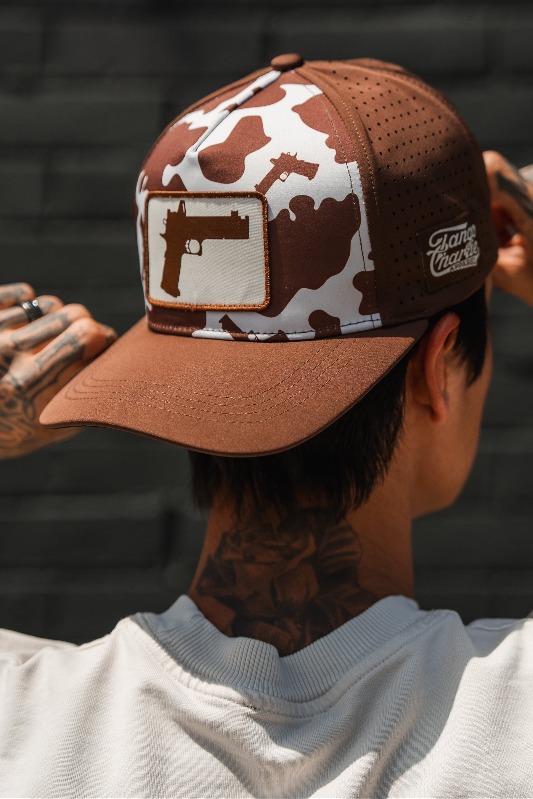 A person with hand and neck tattoos adjusts the Tango Charlie Apparel Moo Moo Pew Hydro snapback hat, featuring a brown and white camouflage pattern, breathable mesh, and gun graphics.