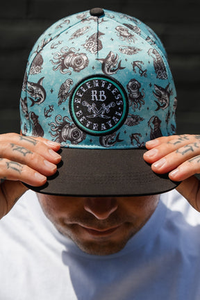 A tattooed person in a white shirt holds the brim of a water-resistant "Sea Swallows the Weak" PVC Snapback by Relentless Betrayal, featuring nautical illustrations and a round branded patch that embodies the pirate lifestyle.