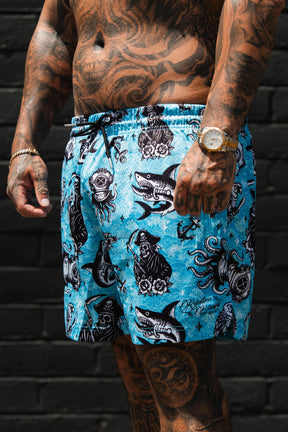 A tattooed man stands before a dark brick wall wearing Relentless Betrayal’s Premium Mesh Shorts | 5.5"- Davy Jones, featuring bold black and white nautical designs—sharks, mermaids, octopuses, and ships—for standout summer style.