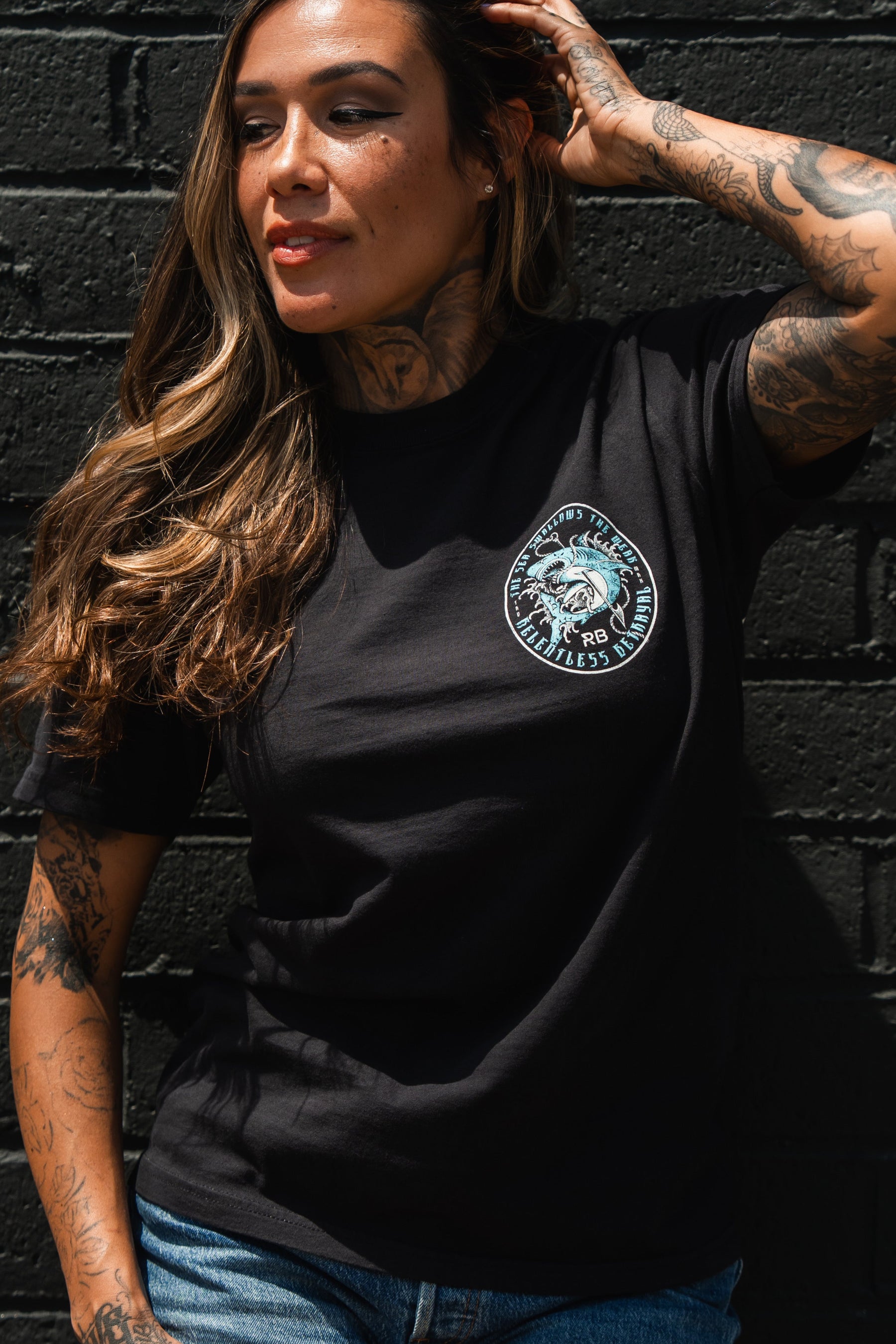 A woman with long hair and tattoos poses against a black brick wall, wearing the Relentless Betrayal "Sea Swallows The Weak" unisex fine jersey cotton t-shirt with denim shorts, looking to the side with one hand on her head.