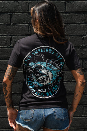 A tattooed person in Relentless Betrayal's Sea Swallows The Weak T-Shirt—featuring a bold shark and ocean waves with circular blue text on the back—and denim shorts stands facing a black brick wall.