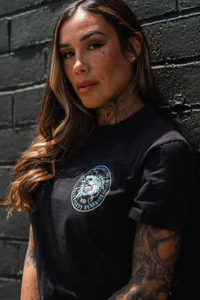 A woman with long brown hair and tattoos leans against a black brick wall, confidently wearing the Relentless Betrayal Sea Swallows The Weak T-Shirt, featuring an eye-catching bold design on fine jersey cotton.