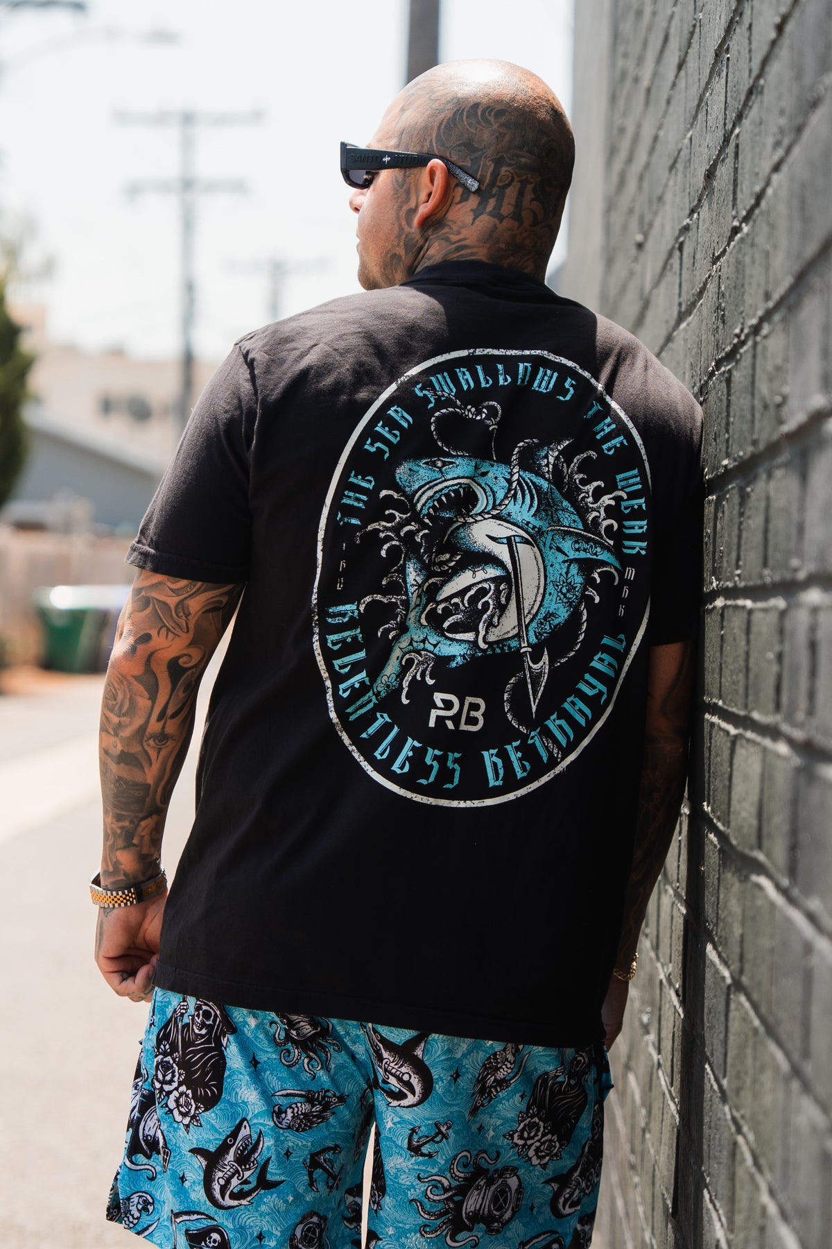 A tattooed man wearing sunglasses stands outside, facing away. He sports the black Sea Swallows The Weak T-Shirt by Relentless Betrayal, styled with blue shark-print shorts in a sunny urban alleyway.