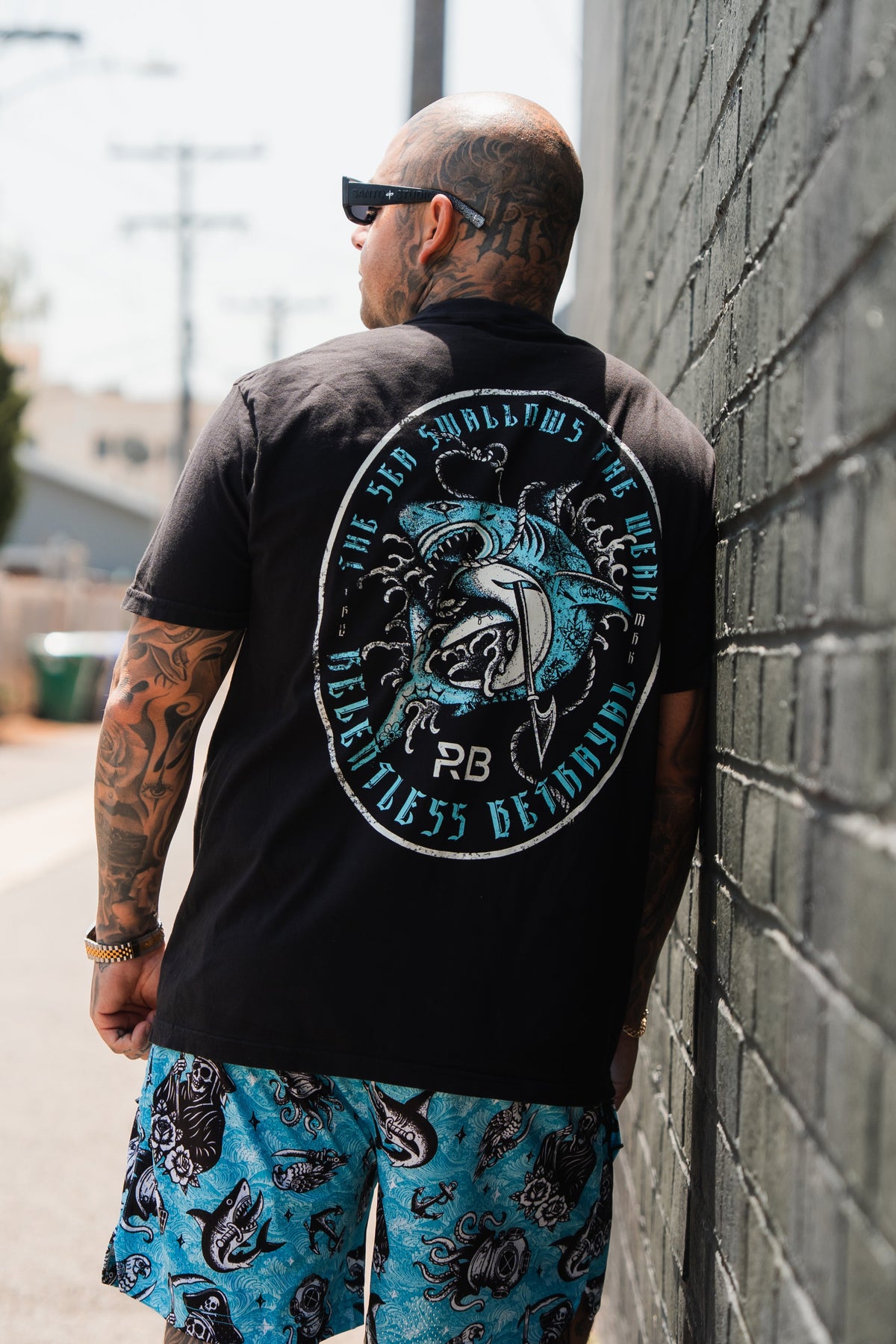 A tattooed man in sunglasses faces a brick wall, wearing a black shark graphic shirt and Relentless Betrayal’s Premium Mesh Shorts | 5.5"- Davy Jones, which feature blue patterns for a bold, active style.