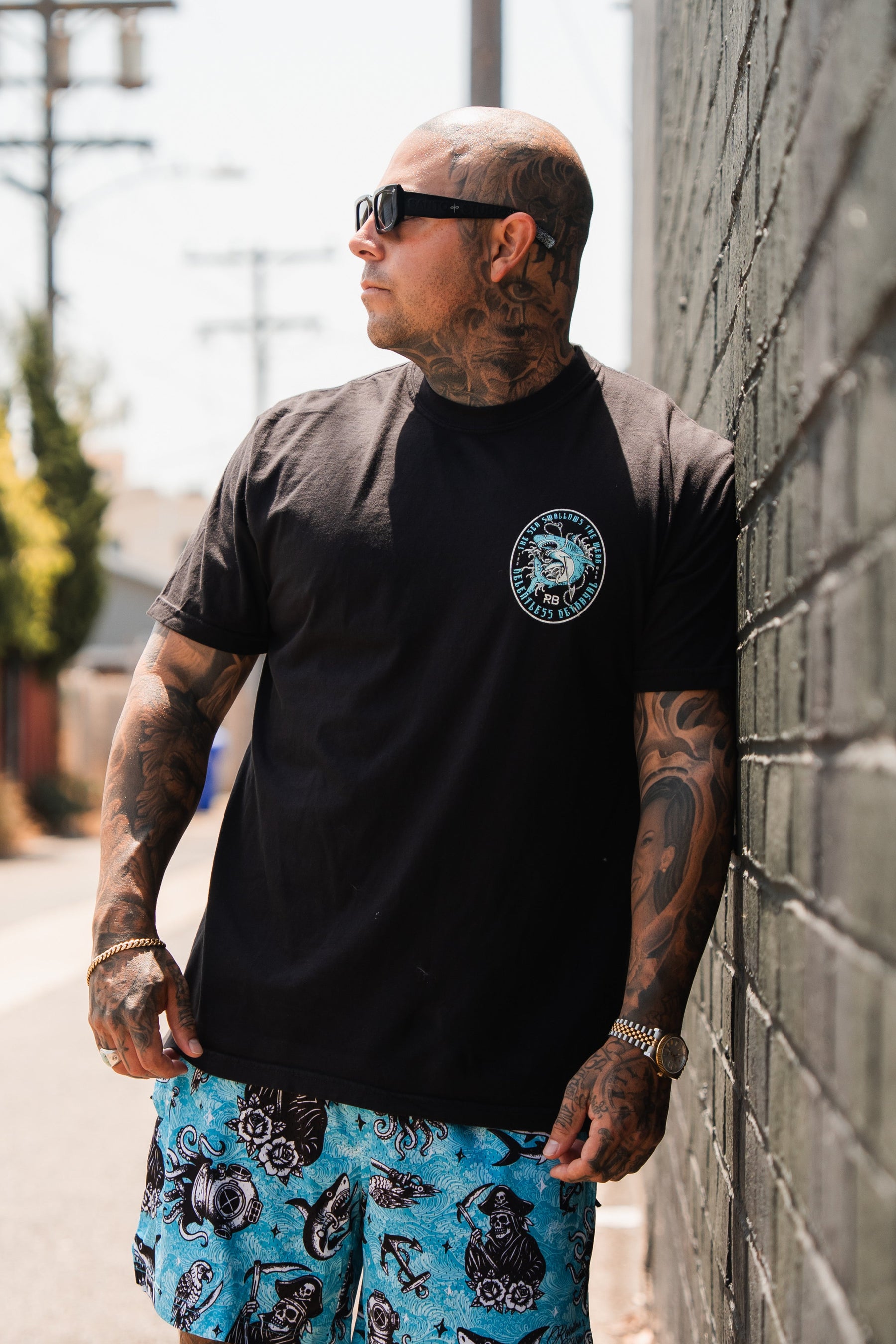 A tattooed man in sunglasses and blue patterned shorts stands against a brick wall outdoors, sporting the Relentless Betrayal Sea Swallows The Weak T-Shirt for a casual fine jersey cotton look.