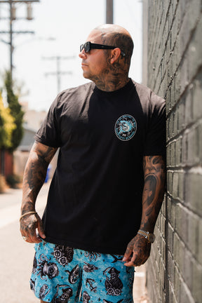A tattooed man in sunglasses and blue patterned shorts stands against a brick wall outdoors, sporting the Relentless Betrayal Sea Swallows The Weak T-Shirt for a casual fine jersey cotton look.