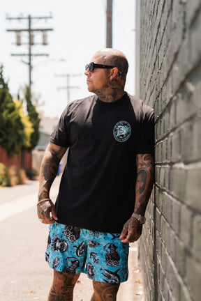 A tattooed man in sunglasses, a black t-shirt, and Relentless Betrayal Premium Mesh Shorts | 5.5" - Davy Jones stands against a brick wall on a sunny day, with power lines and trees in the background.