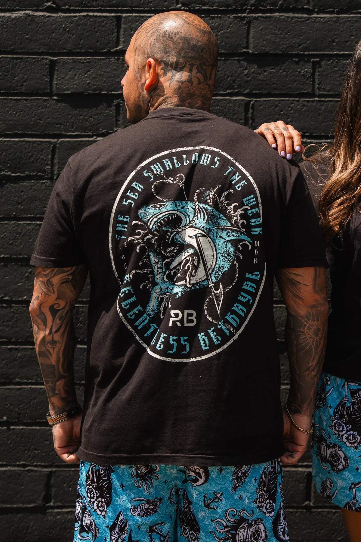A tattooed man wears the Relentless Betrayal Sea Swallows The Weak T-Shirt, featuring a bold bird graphic, with blue patterned shorts as he stands by a black brick wall next to a partially visible companion.
