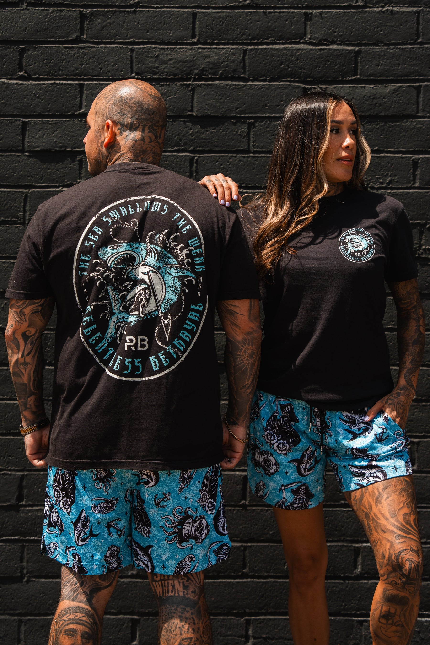 Two tattooed individuals pose against a black brick wall, confidently wearing matching Relentless Betrayal Sea Swallows The Weak T-Shirts, each featuring a bold back design, paired with blue shorts patterned in black for a standout look.