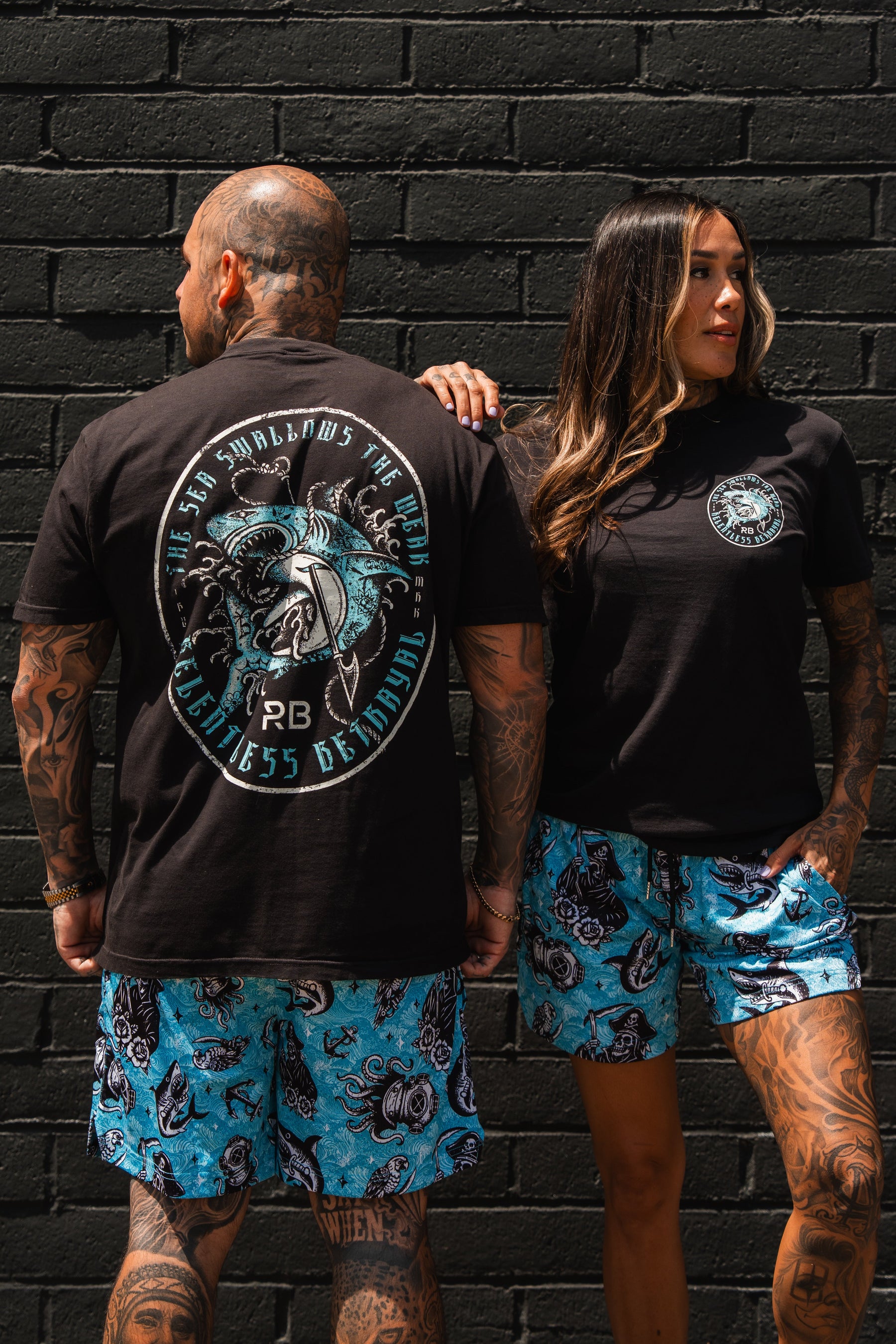 Two people wearing matching black t-shirts and blue shorts with a design against a black brick wall.