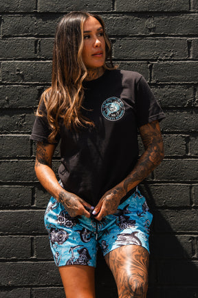 A woman with long brown hair and tattoos stands against a black brick wall, wearing the Relentless Betrayal Sea Swallows The Weak T-Shirt and bold blue shorts. She gazes to the side with a relaxed expression.