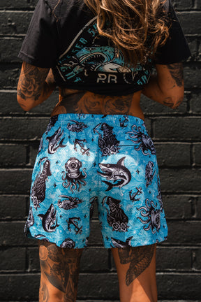 A tattooed person stands against a black brick wall wearing a black graphic tee and Relentless Betrayal Premium Mesh Shorts | 5.5"- Davy Jones, featuring bold, black-and-white sea creature designs in bright blue.
