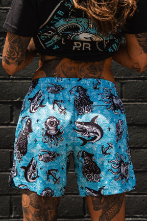 A tattooed individual wears Relentless Betrayal Premium Mesh Shorts | 5.5"- Davy Jones, featuring black and white nautical designs—sharks, divers, octopuses, anchors, and ship wheels—while standing by a dark brick wall.