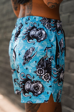 A person stands near a gray brick wall wearing Relentless Betrayal's Premium Mesh Shorts | 5.5"–Davy Jones, featuring bright blue fabric with black and white nautical graphics like diving helmets, birds, and octopuses.