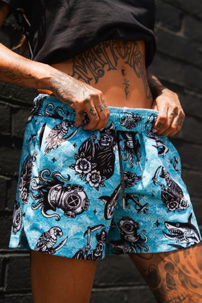 A tattooed person stands by a dark brick wall wearing Relentless Betrayal’s Premium Mesh Shorts | 5.5"–Davy Jones, featuring bold black and white nautical patterns—octopuses, sharks, divers, and anchors—for any unisex athletic style.