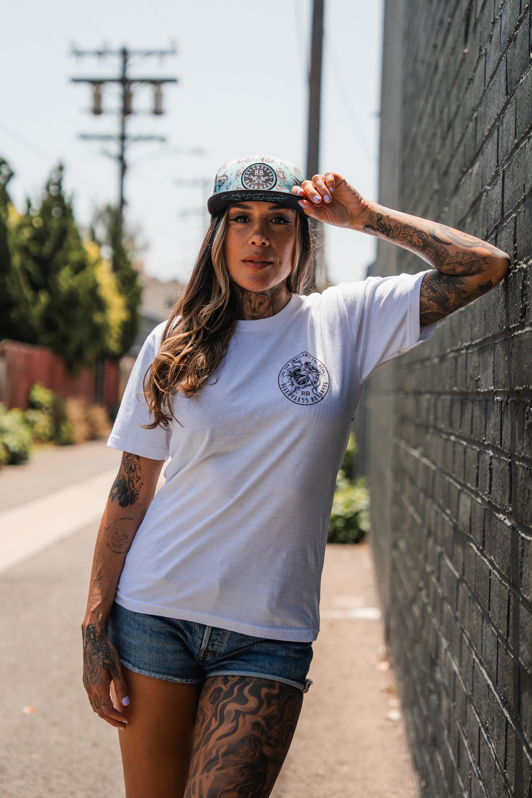 A tattooed woman in the Relentless Betrayal "Sea Swallows The Weak" white T-shirt, denim shorts, and a cap stands against a brick wall on a sunny day, holding her cap's brim and looking at the camera.
