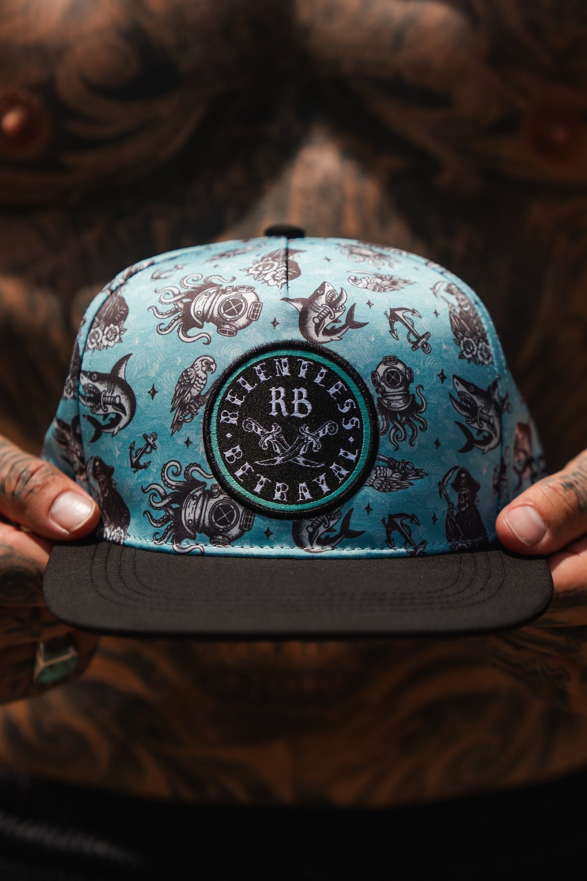A tattooed person holds the Relentless Betrayal Sea Swallows the Weak PVC Snapback, a turquoise and black hat with tattoo-inspired illustrations and a bold round patch.
